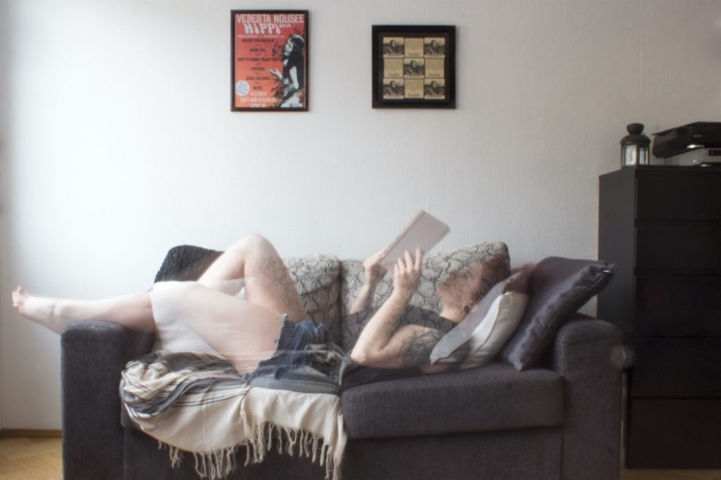 Nainen makaa sohvalla kirja kädessään. Taustalla valkoinen seinä, jossa on kaksi julistetta. Nainen on kuvassa läpikuultava.