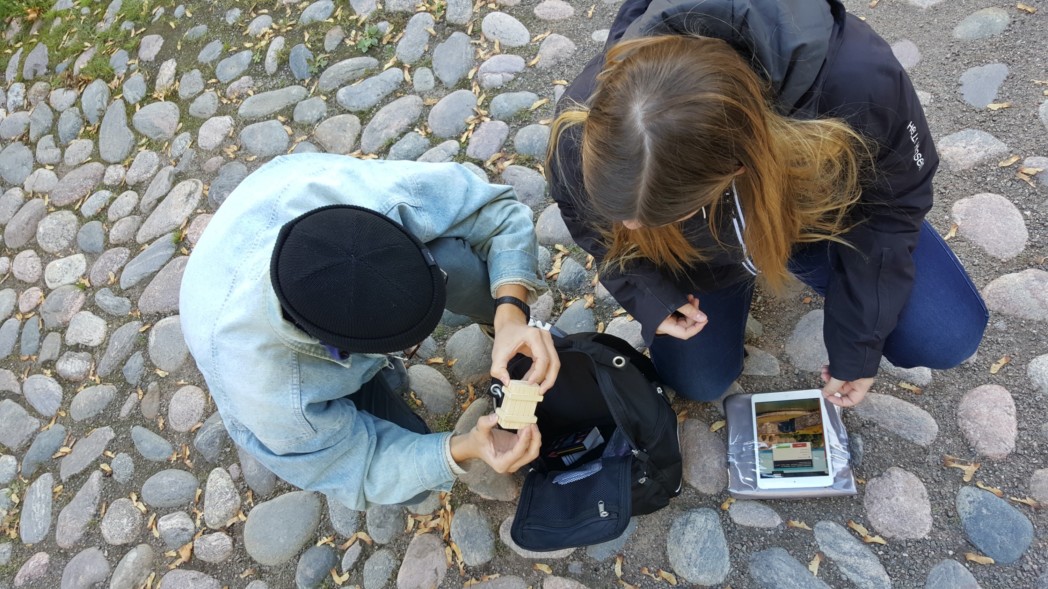 kaksi nuorta henkilöä tutkii esinettä mukulakivetyksellä, kuva on otettu yläsuunnasta. Maassa on älylaite.